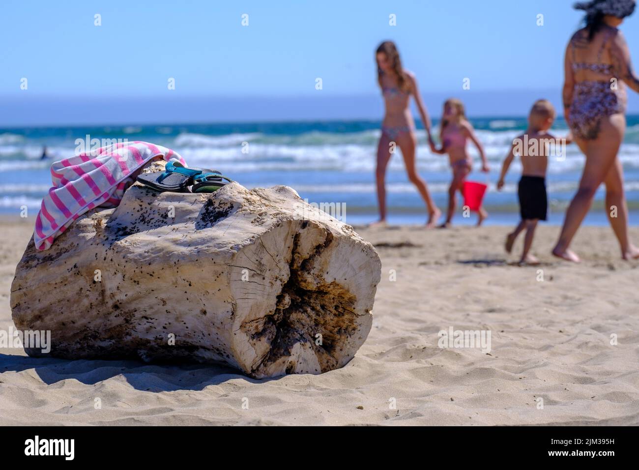 Close-up of a log on the beach with a towel & flip flops on top. People walking on the beach & the ocean in the background. Stock Photo