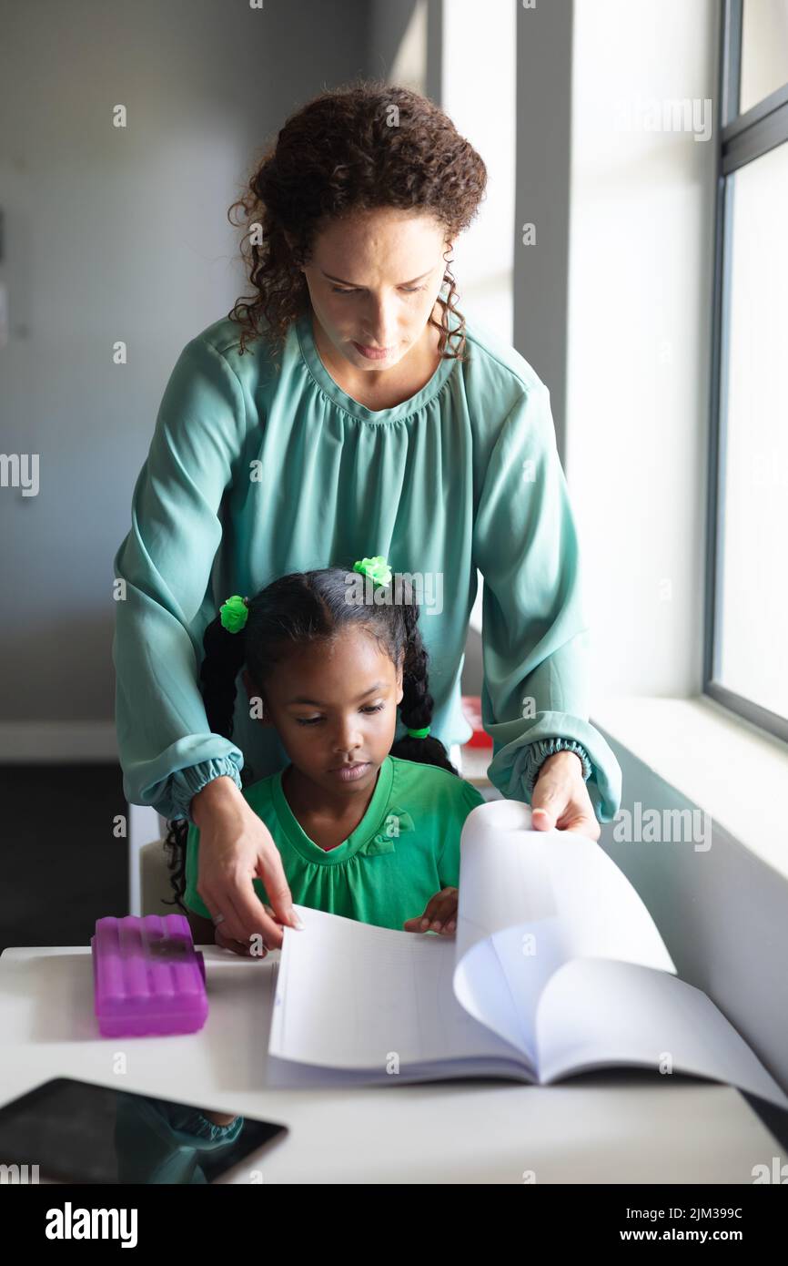 Caucasian young female teacher assisting african american schoolgirl studying at desk in classroom. unaltered, education, childhood, learning, teachin Stock Photo