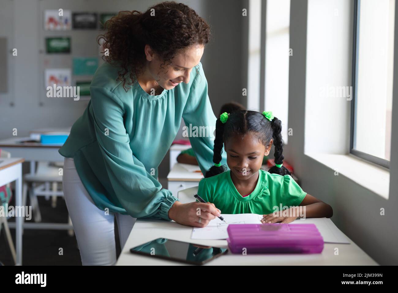 Caucasian young female teacher assisting african american elementary schoolgirl writing on book Stock Photo