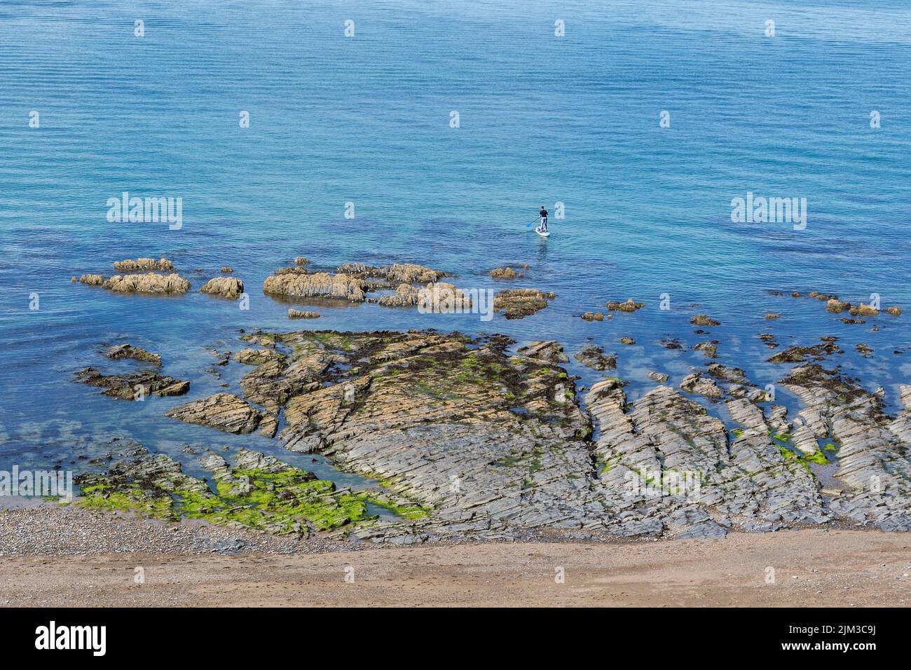 A man paddle boarding in the sea by a rocky shoreline. Health, fitness,holiday, recreation, solitude concept. Stock Photo