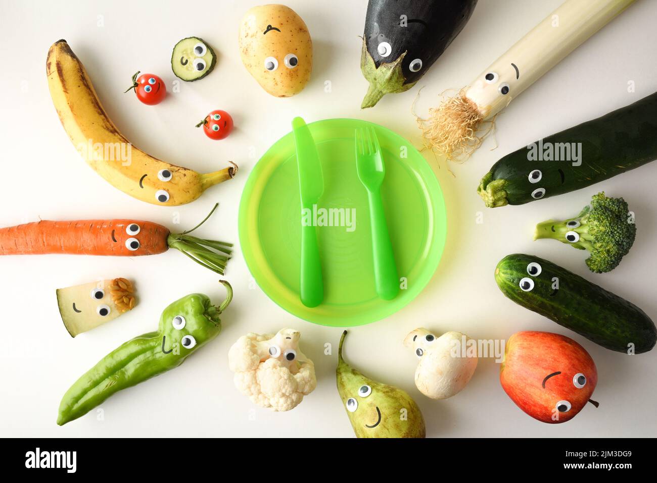 Backgound with attractive plate of funny fruits and vegetables served for child with red children's cutlery on white table. Top view. Stock Photo