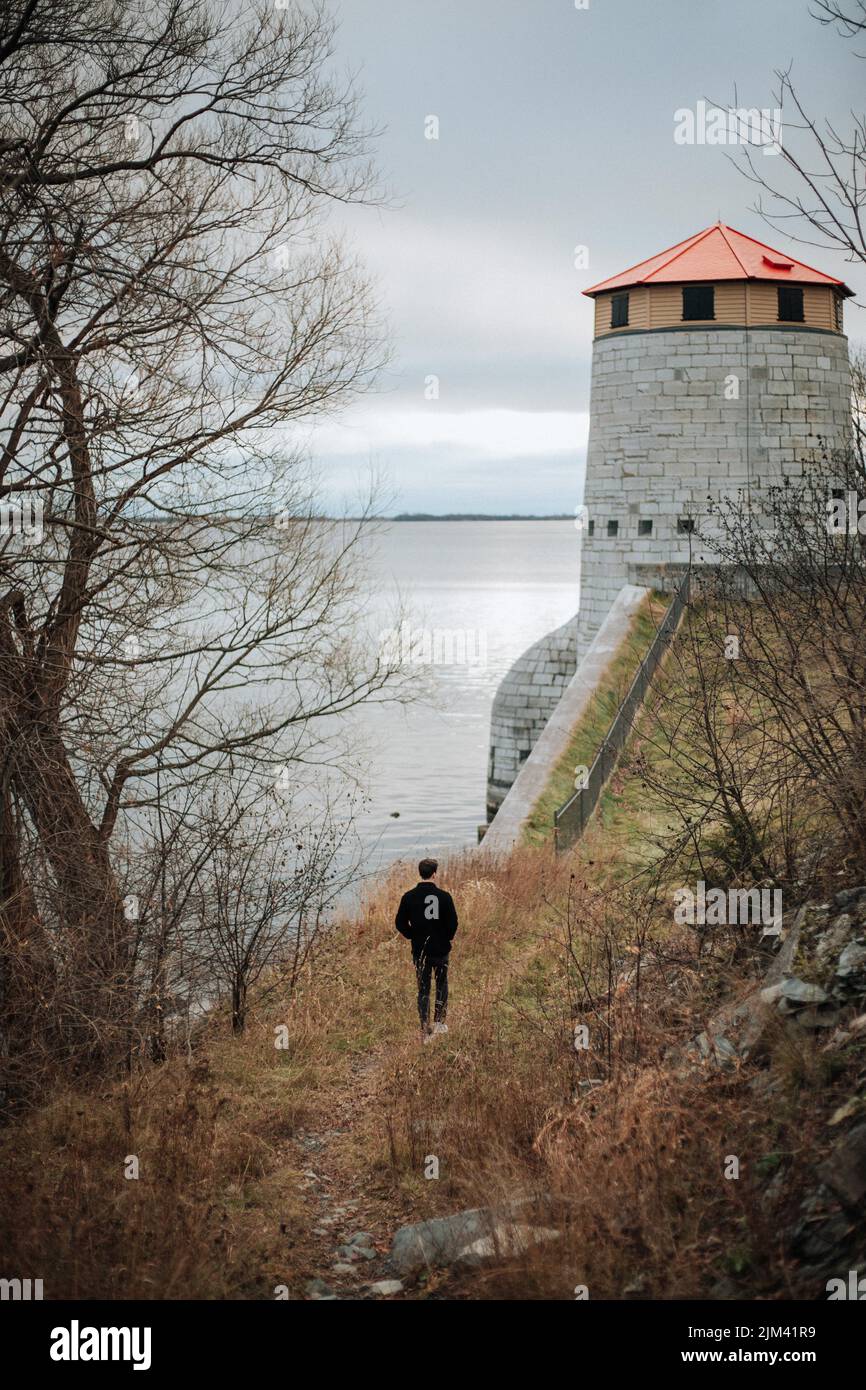 A back shot of a young man walking on unpaved road to the Murney Tower National Historic Site of Canada during daytime in Kingston, Ontario, Canada Stock Photo
