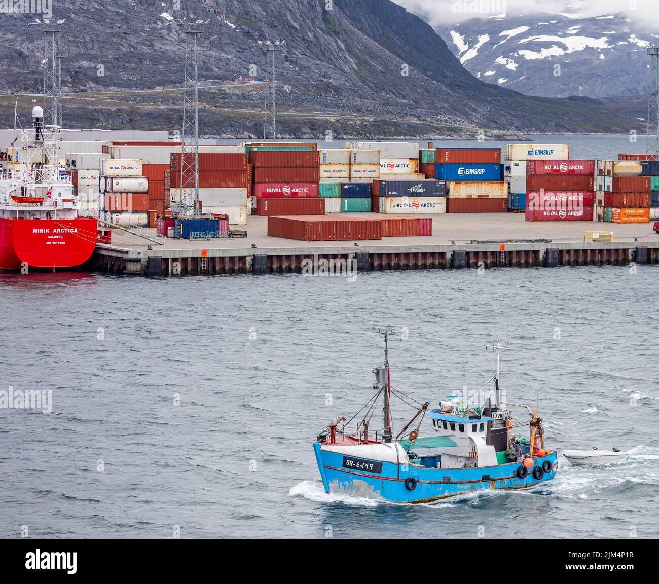Small fishing returning to port in front of containers stacked up at the port in Nuuk with snow capped mountains in the background in Nuuk, Greenland Stock Photo