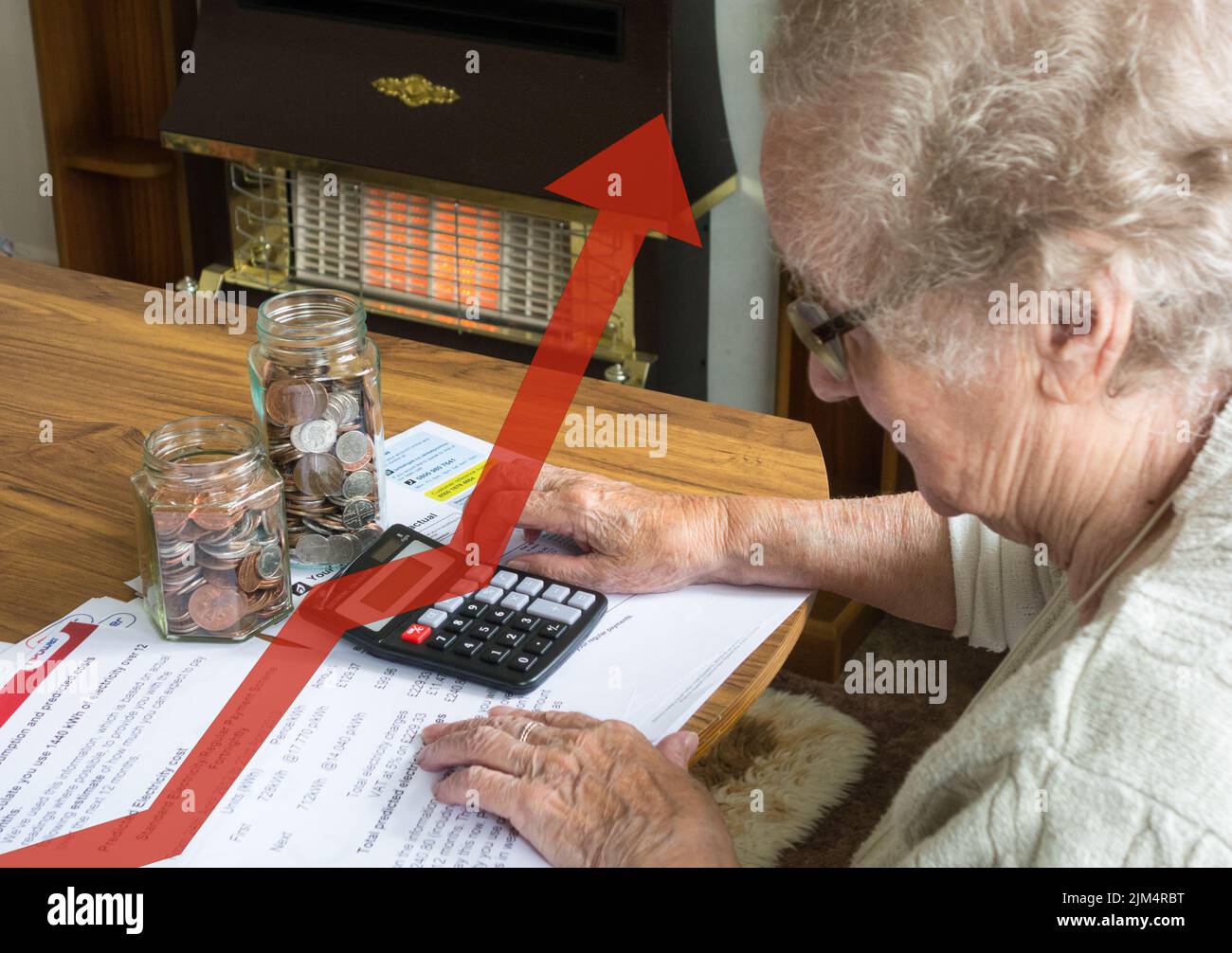 Elderly lady in her nineties looking at gas, electric, utility bills with calculator and jars of coins on table. Gas fire on in background. Stock Photo