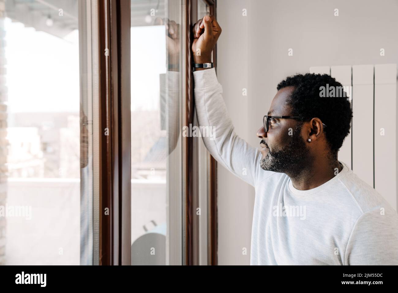Black bearded man wearing eyeglasses looking in window at home Stock Photo