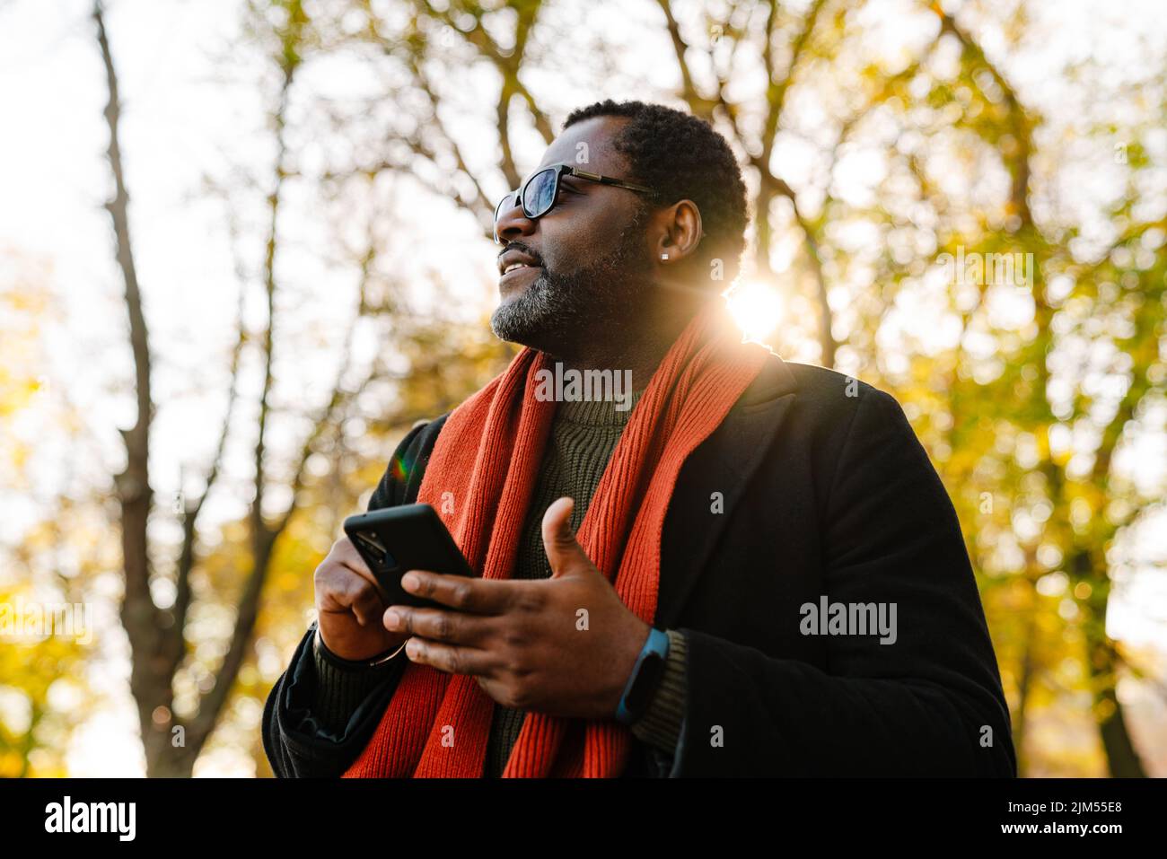 Black bearded man in sunglasses using cellphone while strolling at park Stock Photo