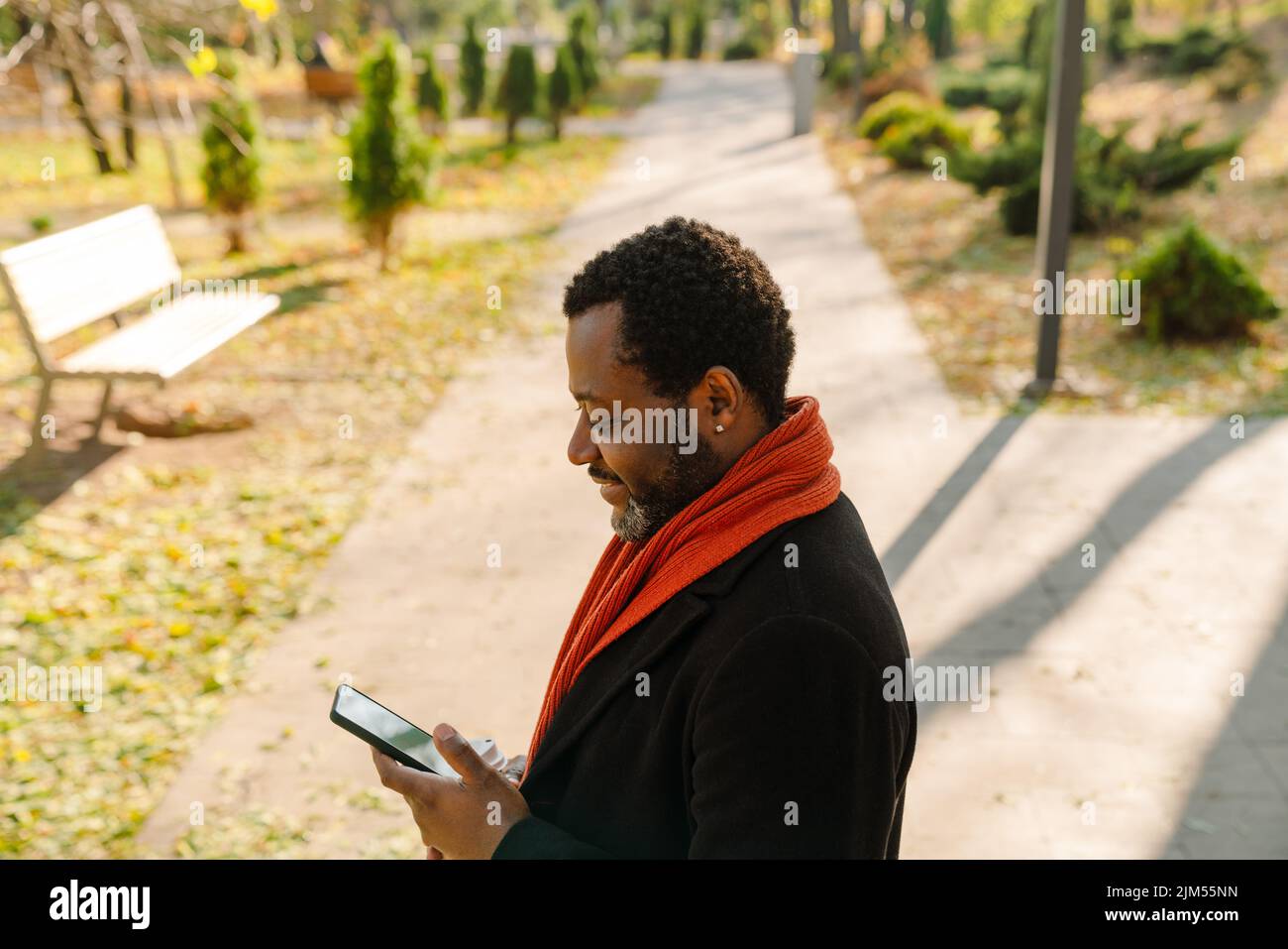Black bearded man using cellphone and drinking coffee while strolling in park Stock Photo