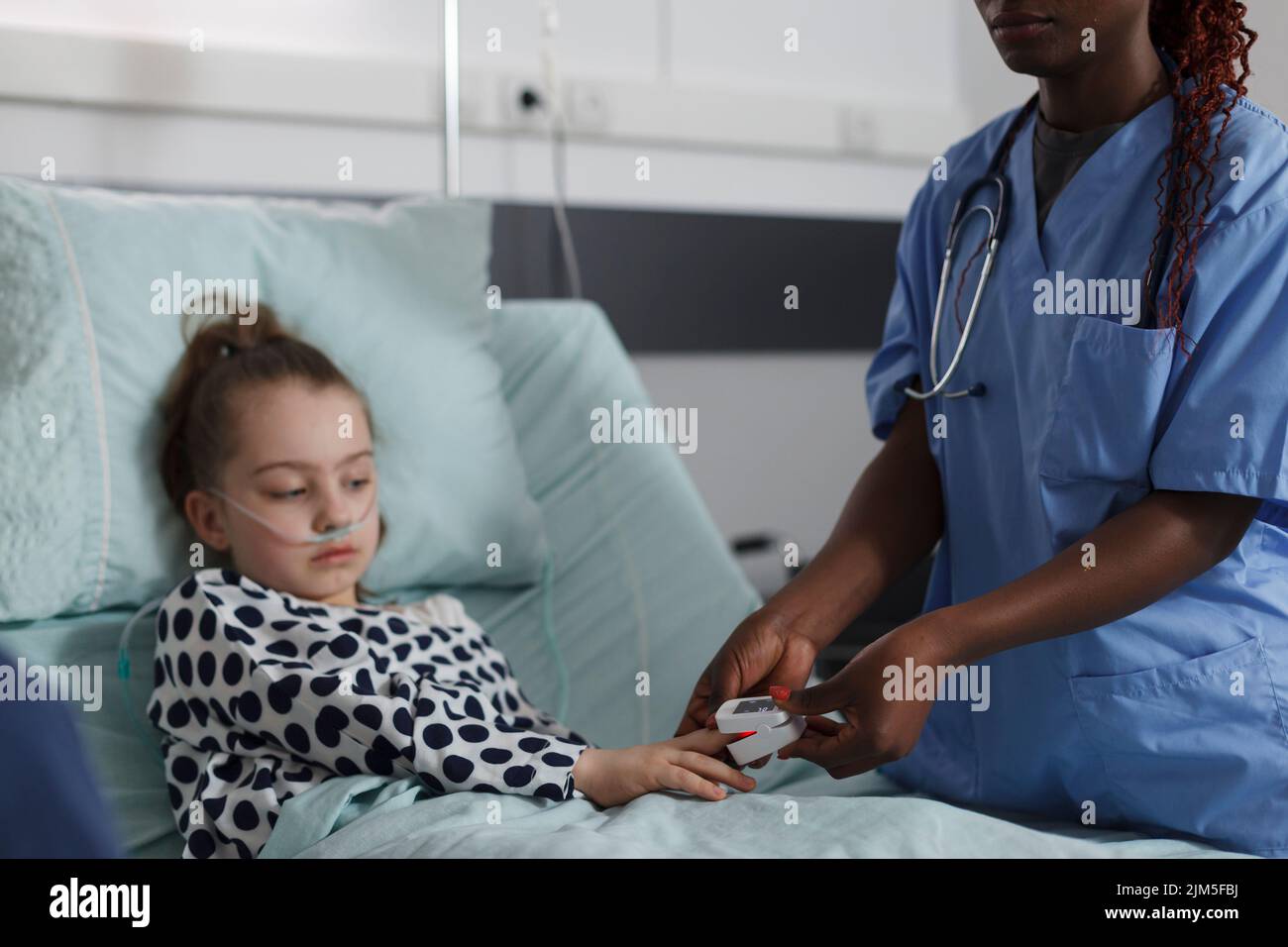 Ill kid sitting in hospital pediatric ward while medical staff monitoring health condition using oximeter. Nurse measuring oxygen levels of hospitalized sick little girl resting on patient bed Stock Photo