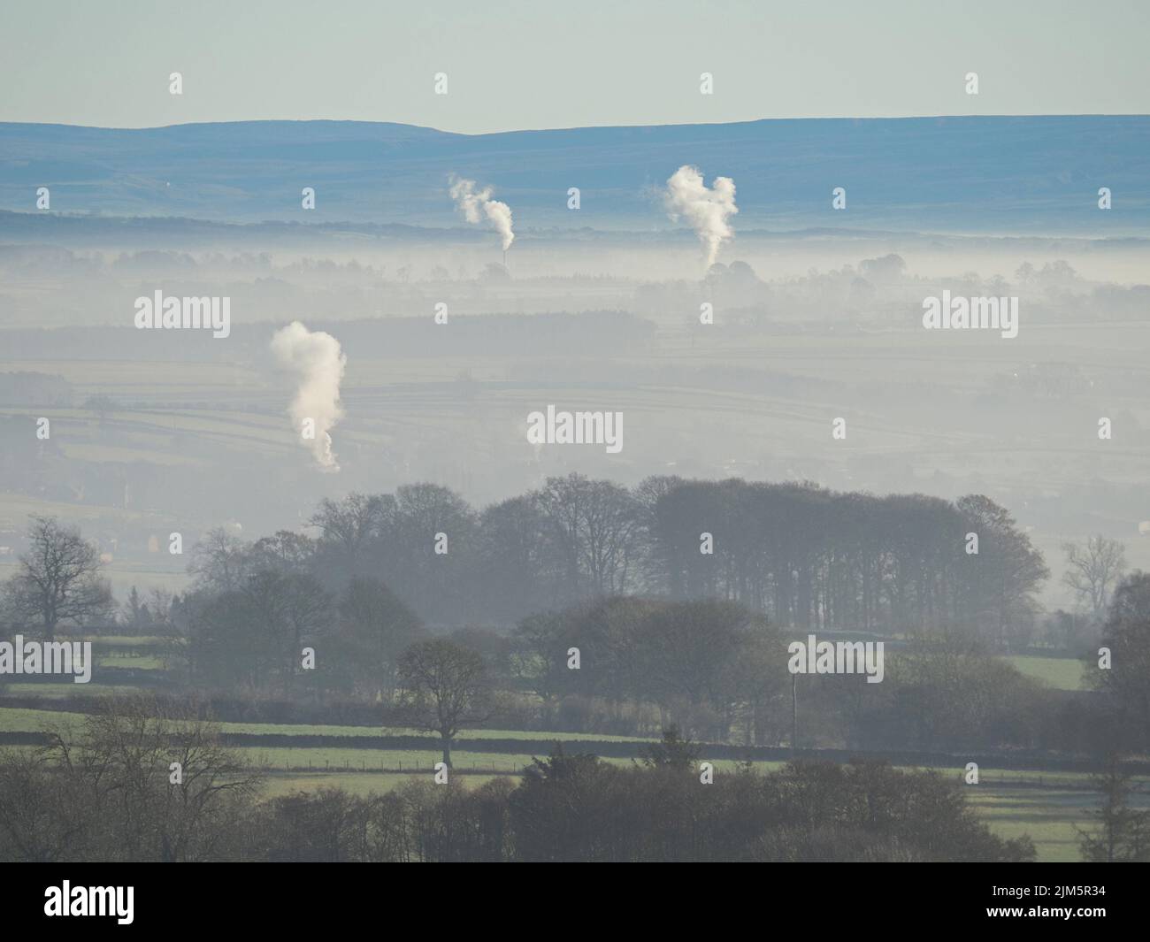 columns of white smoke rise above a veil of mist in valley below Northern Pennines on still frosty morning of low Winter sunshine - Cumbria,England,UK Stock Photo