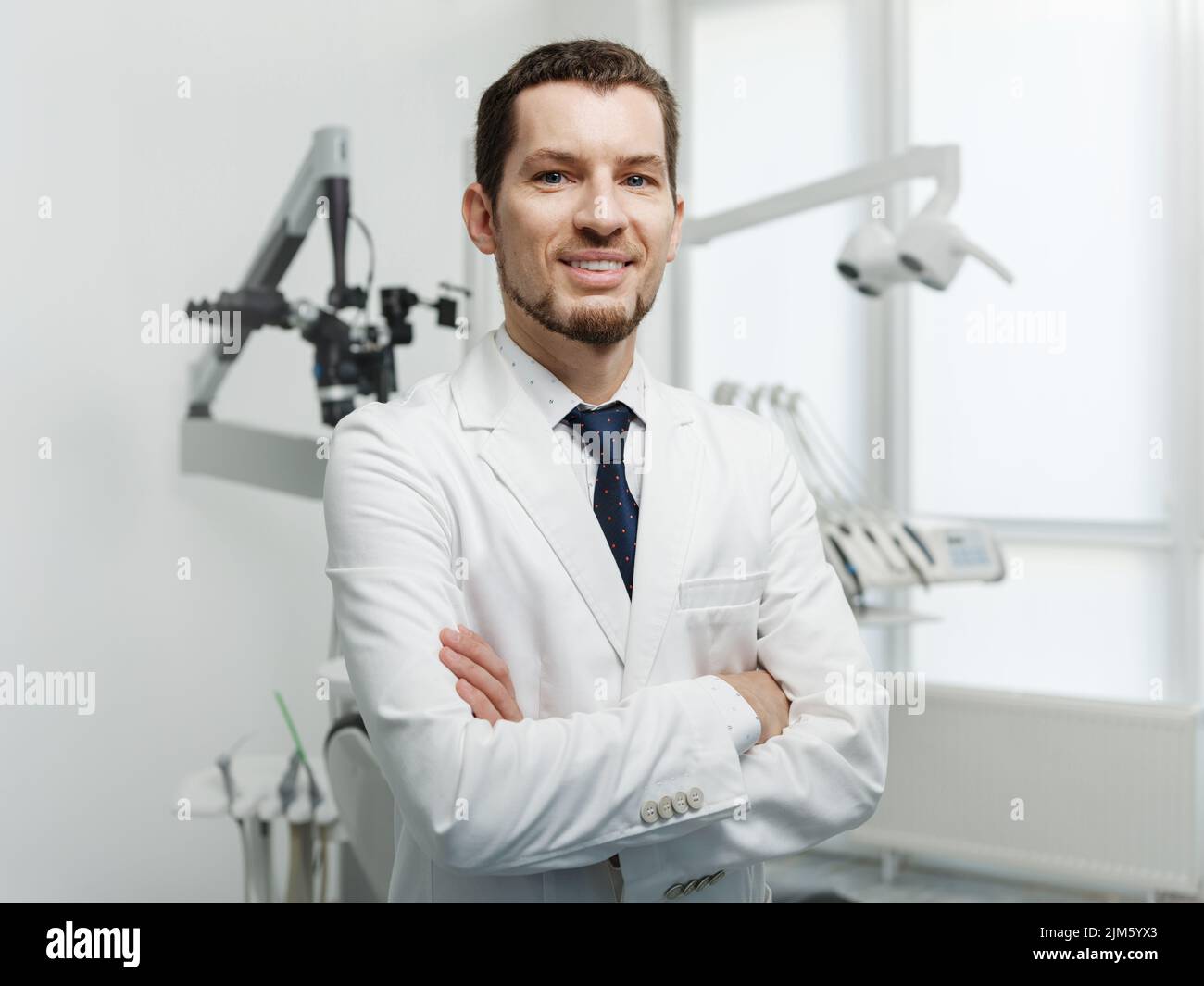Happy male dentist in white lab coat posing with hands crossed and