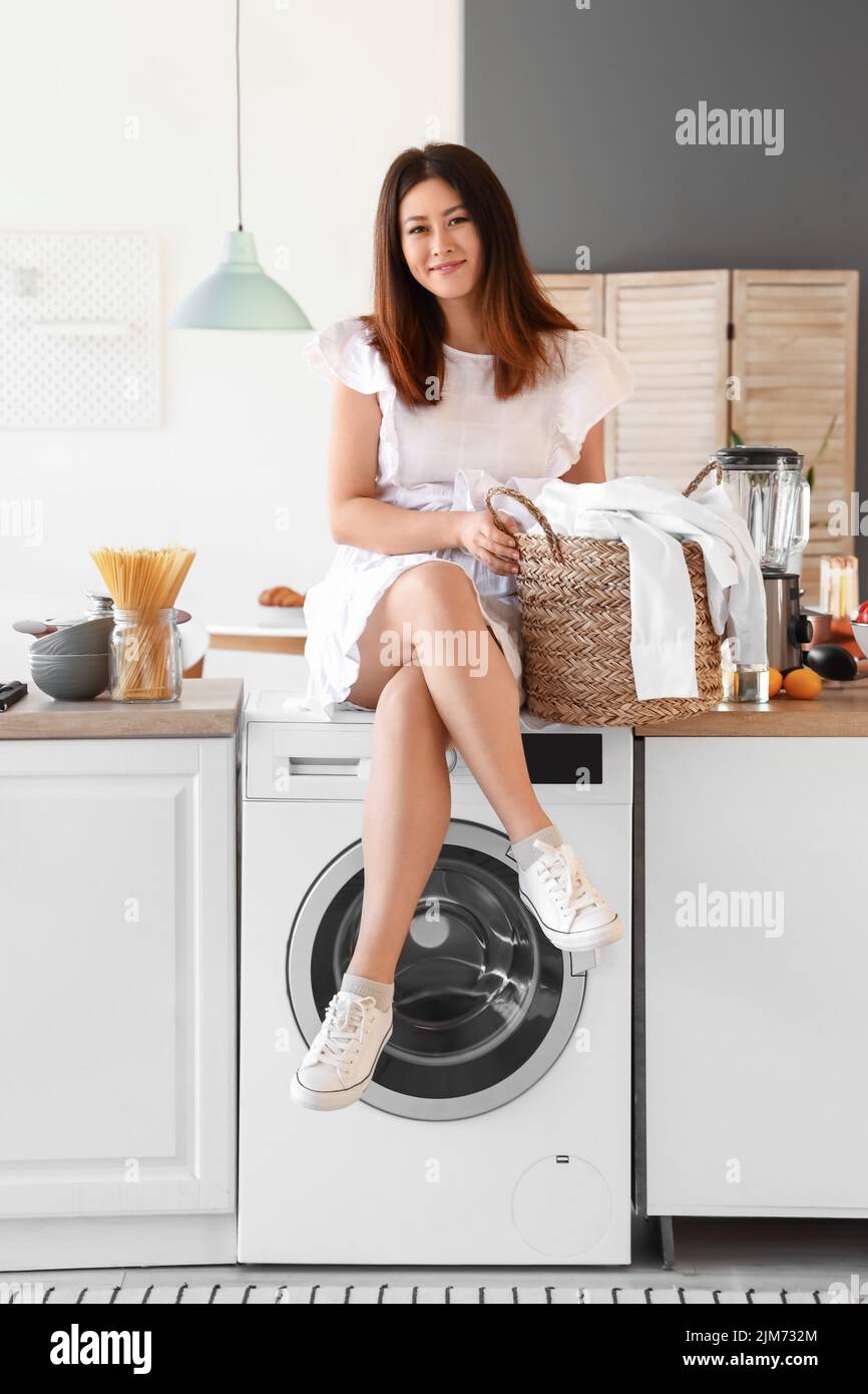 Young Asian woman with laundry basket sitting on washing machine in ...