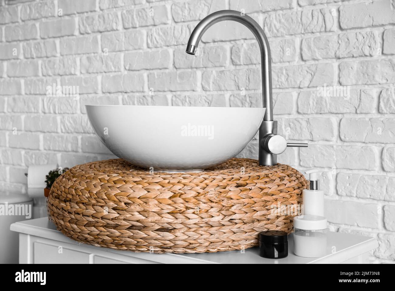 Table with sink, rattan pouf and cosmetic products near white brick wall Stock Photo