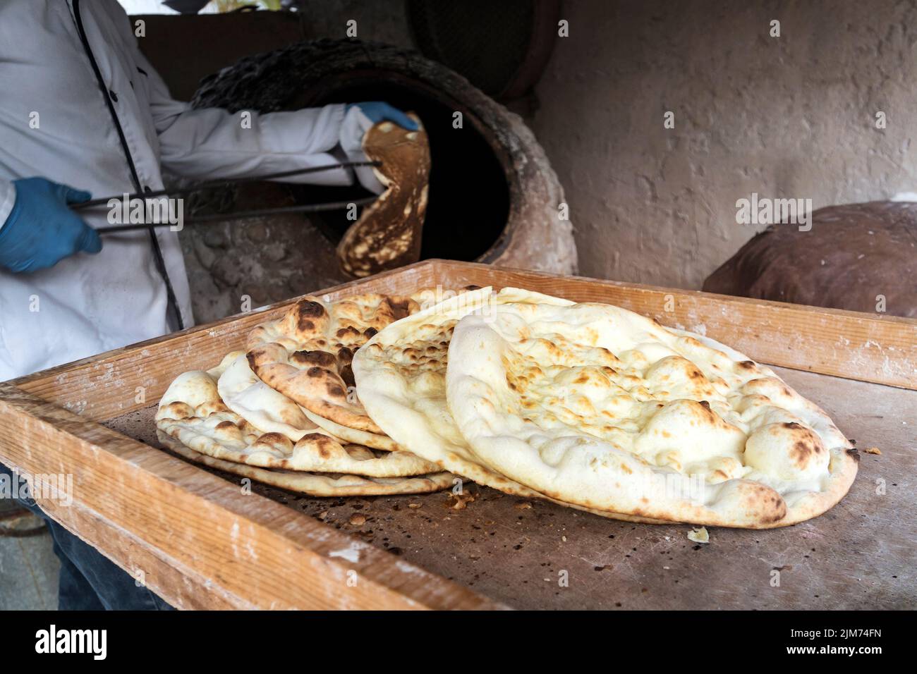 aged man makes traditional bread in old round stone oven in rural village. Large flat freshly baked tortillas lie on the table. Hot bread. Central Asi Stock Photo