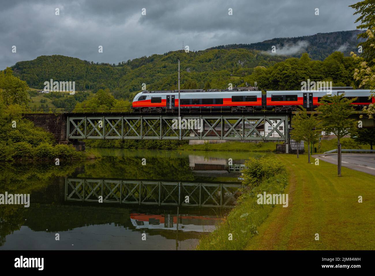 Modern train running over metal trestle bridge at Reichraming, Austria. Nice reflection in the water below. Cloudy day but with lots of color. Stock Photo