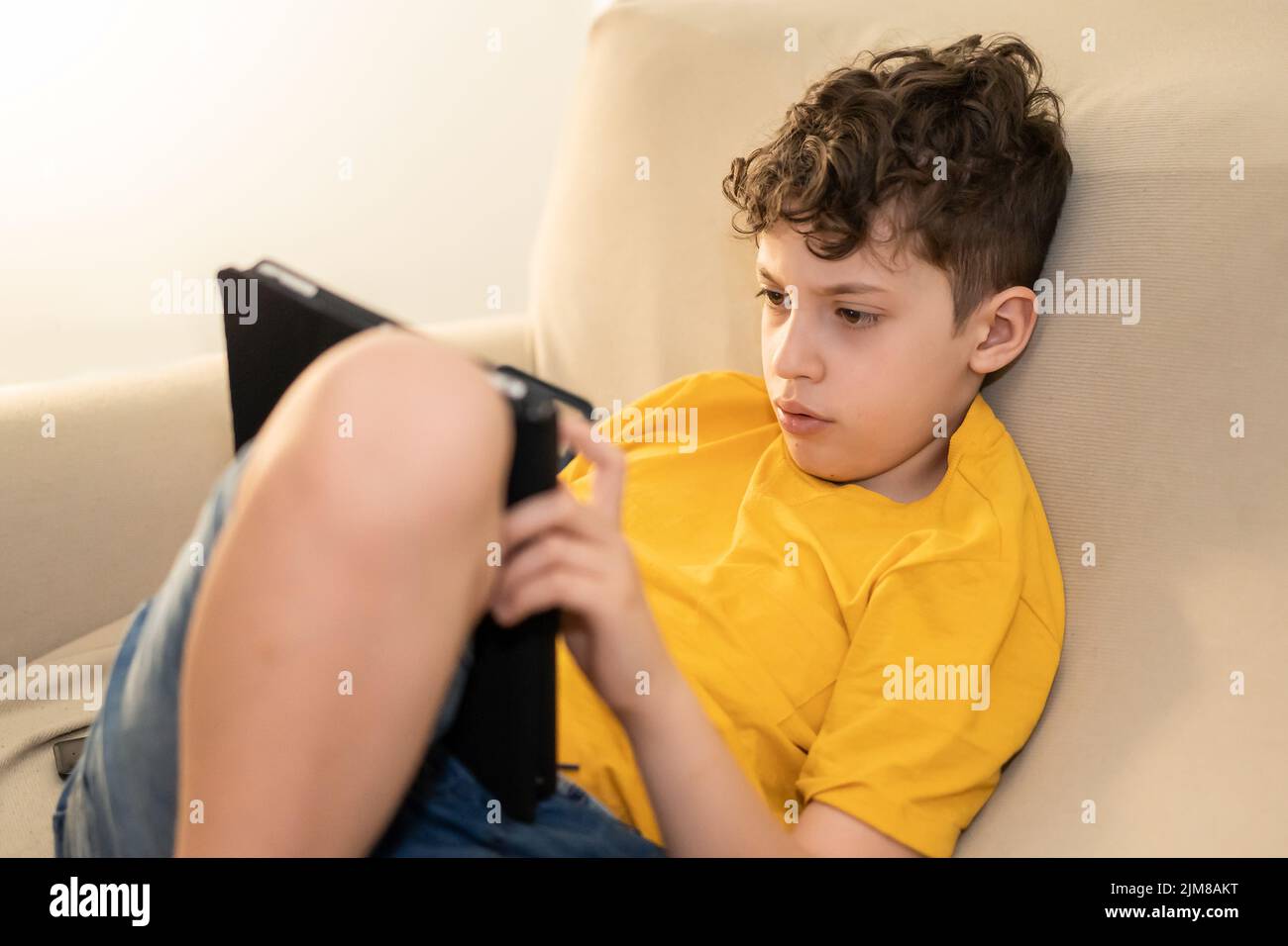 School aged kid using a tablet relaxing on the coach indoor. Children addicted from technology studying online from home - education, addiction, peopl Stock Photo