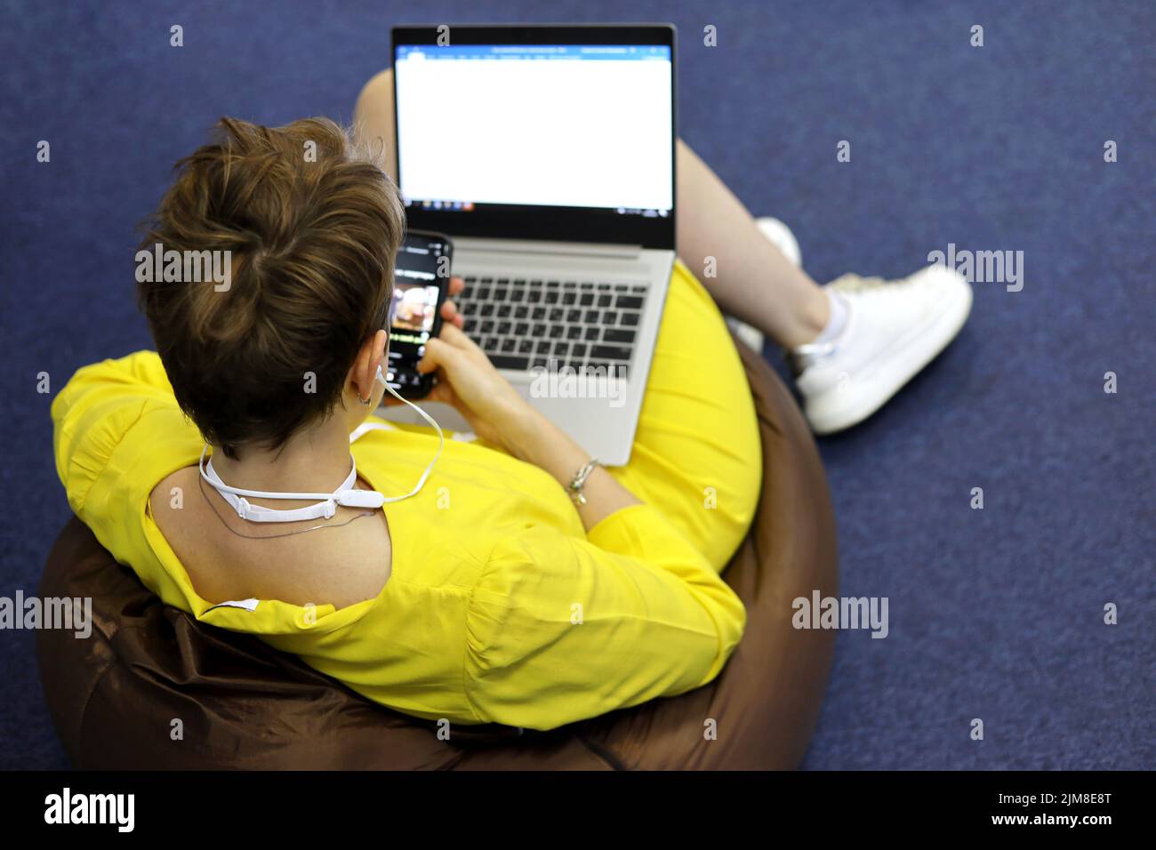 Woman in headphones wearing yellow dress sitting on padded stool with smartphone in hand and laptop on her knees, top view. Listening music Stock Photo