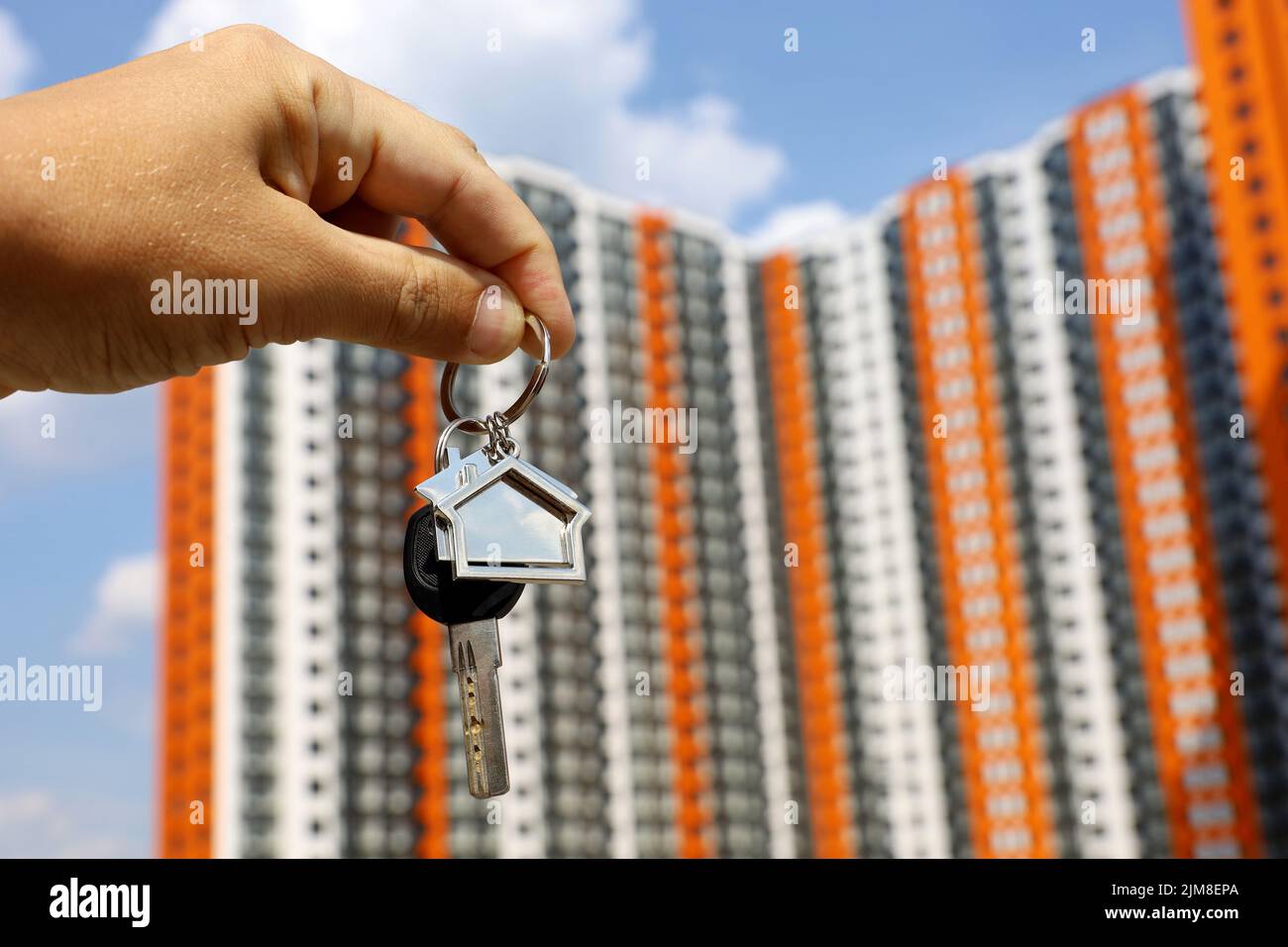 Real estate agent, keychain in shape of a house and key in male hand on background of new buildings. Byung apartment or renting property in summer Stock Photo
