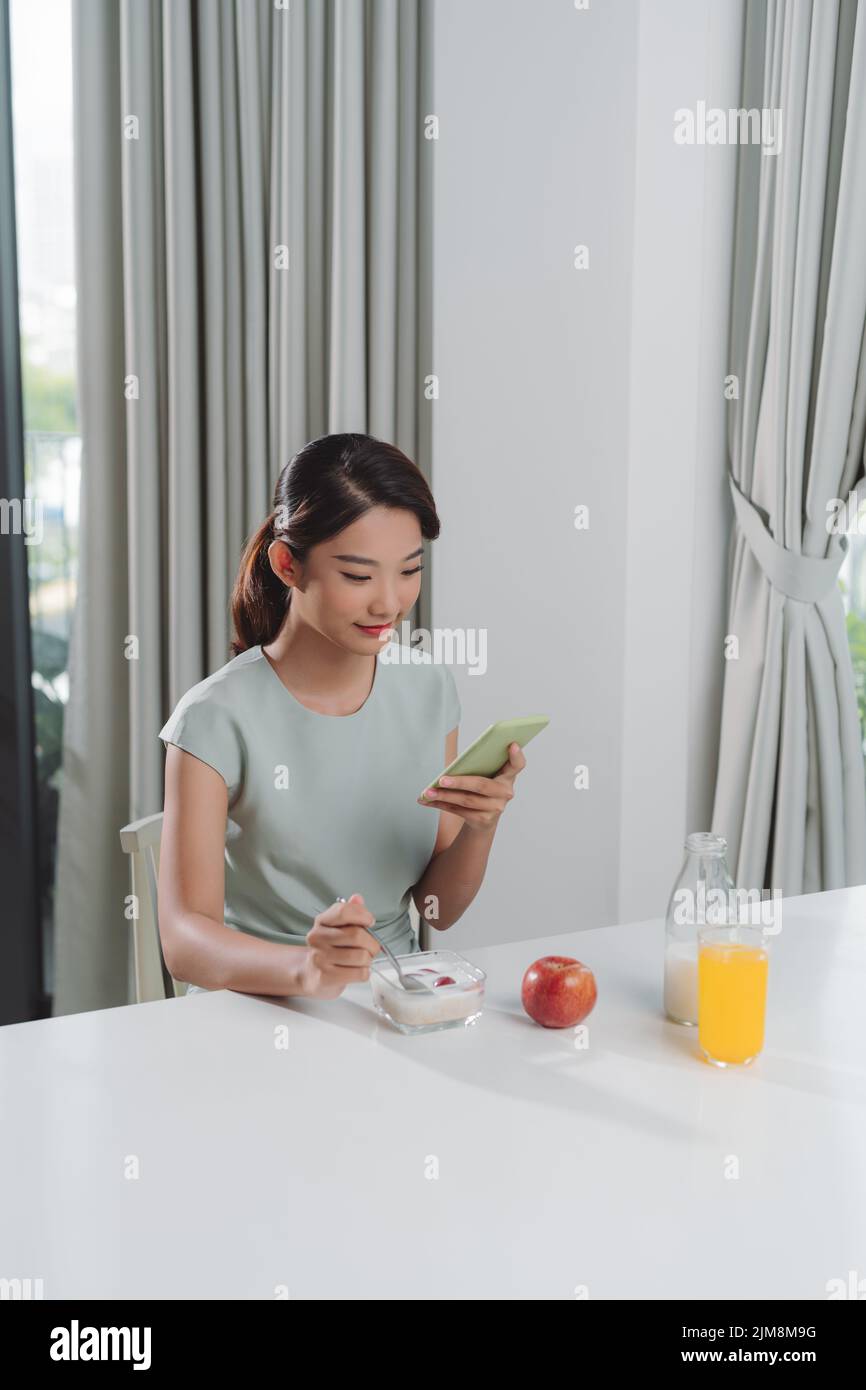 Beautiful mature woman eating oat and milk while sending messages with smartphone at home. Stock Photo