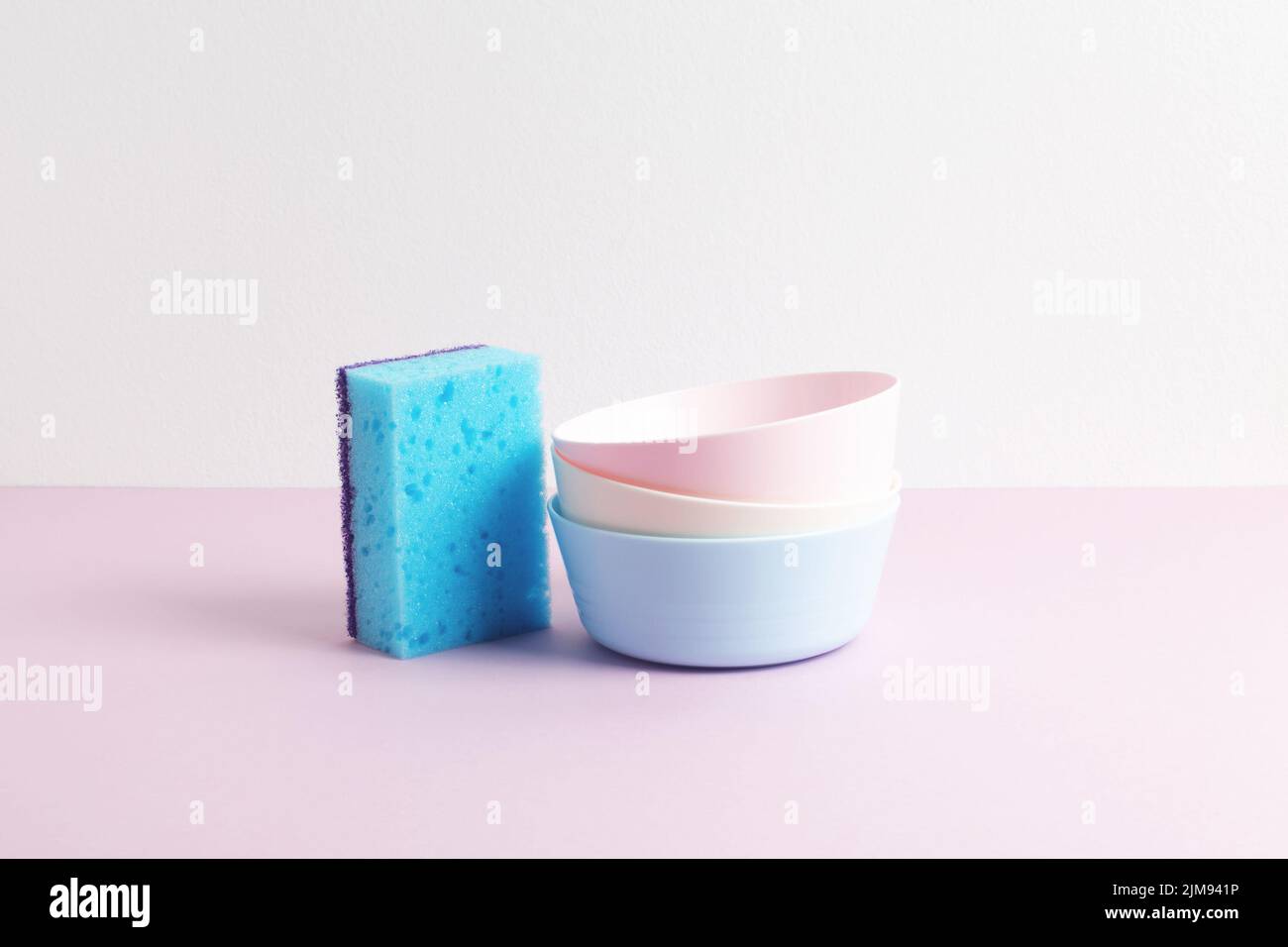 Blue sponge and colorful clean plates on pink and white background. Pastel colors. Front view. Dishwashing concept. Stock Photo