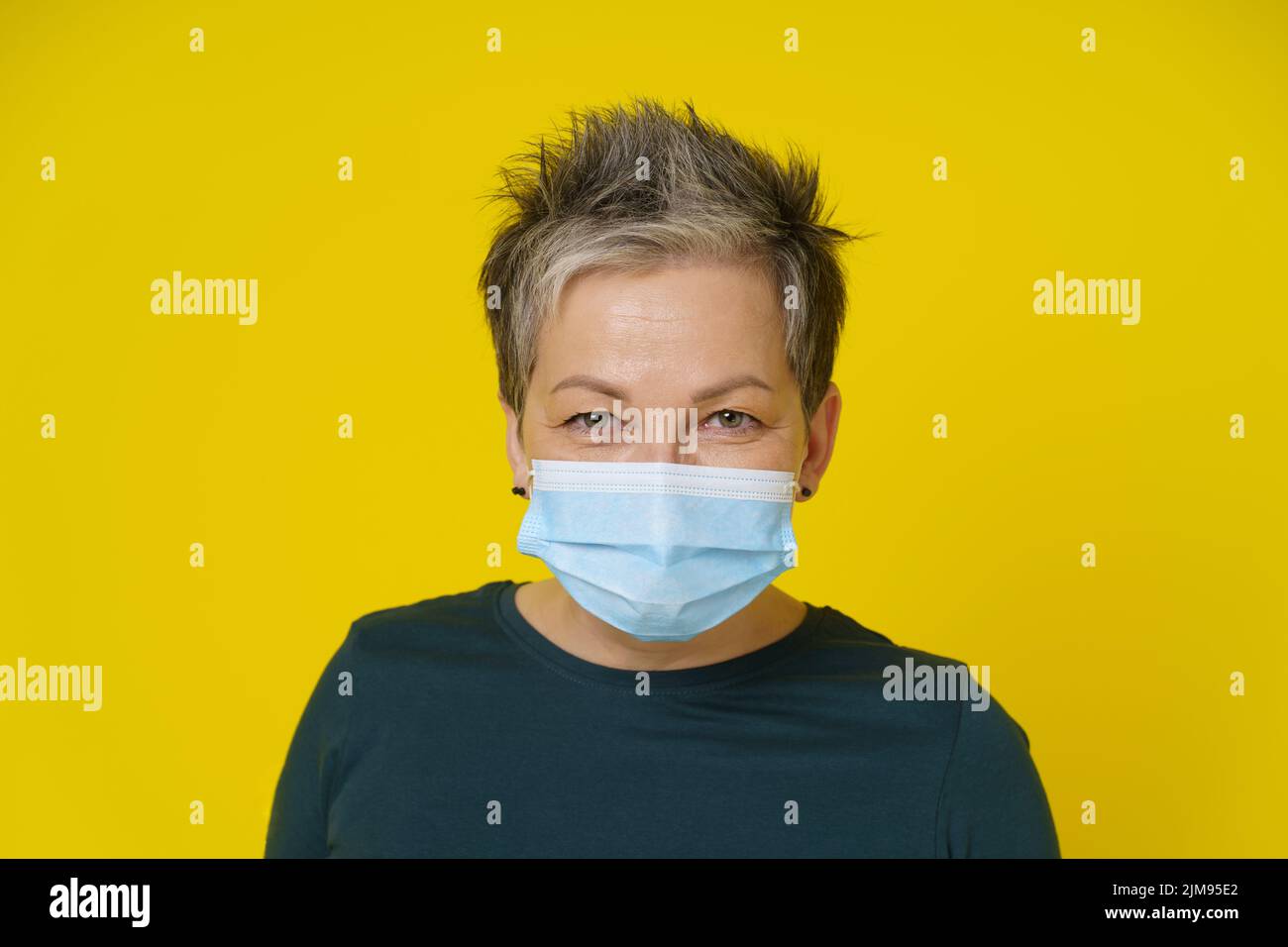 Mature woman wearing medical face mask with happy eyes smiling looking at camera wearing green blouse isolated on yellow background. Close up adult woman in medical mask.  Stock Photo