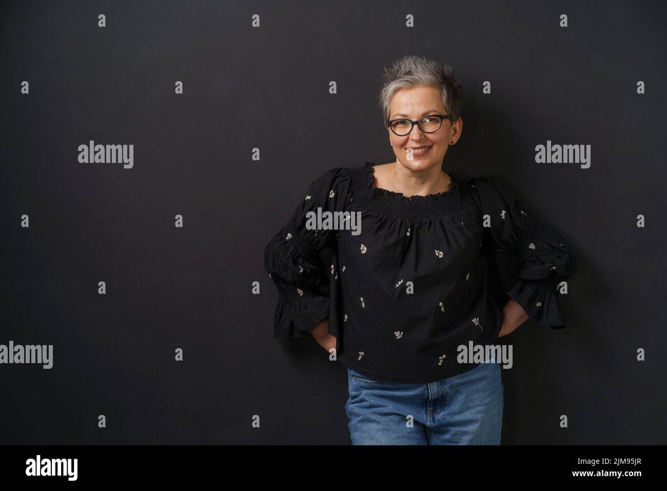 Gorgeous mature grey haired businesswoman with grey hair leaned on wall isolated on black background. Elegant woman with short hair. Image of successful mature woman. Aged beauty. Stock Photo
