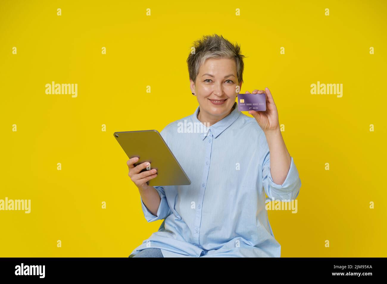 Mature woman with grey hair holding credit, debit card and tablet pc in hand making online payment or shopping online, isolated on yellow background. E-commerce, online banking concept. Stock Photo