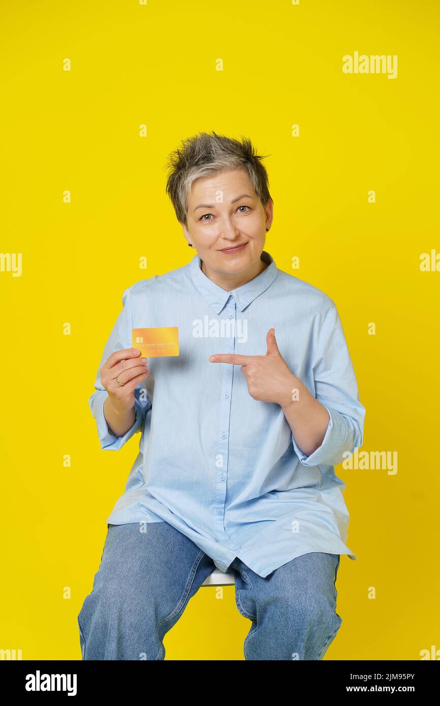 Grey haired mature woman holding credit, debit card in hand pointing at it making online payment or shopping online, isolated on yellow background. E-commerce, online banking concept. Stock Photo