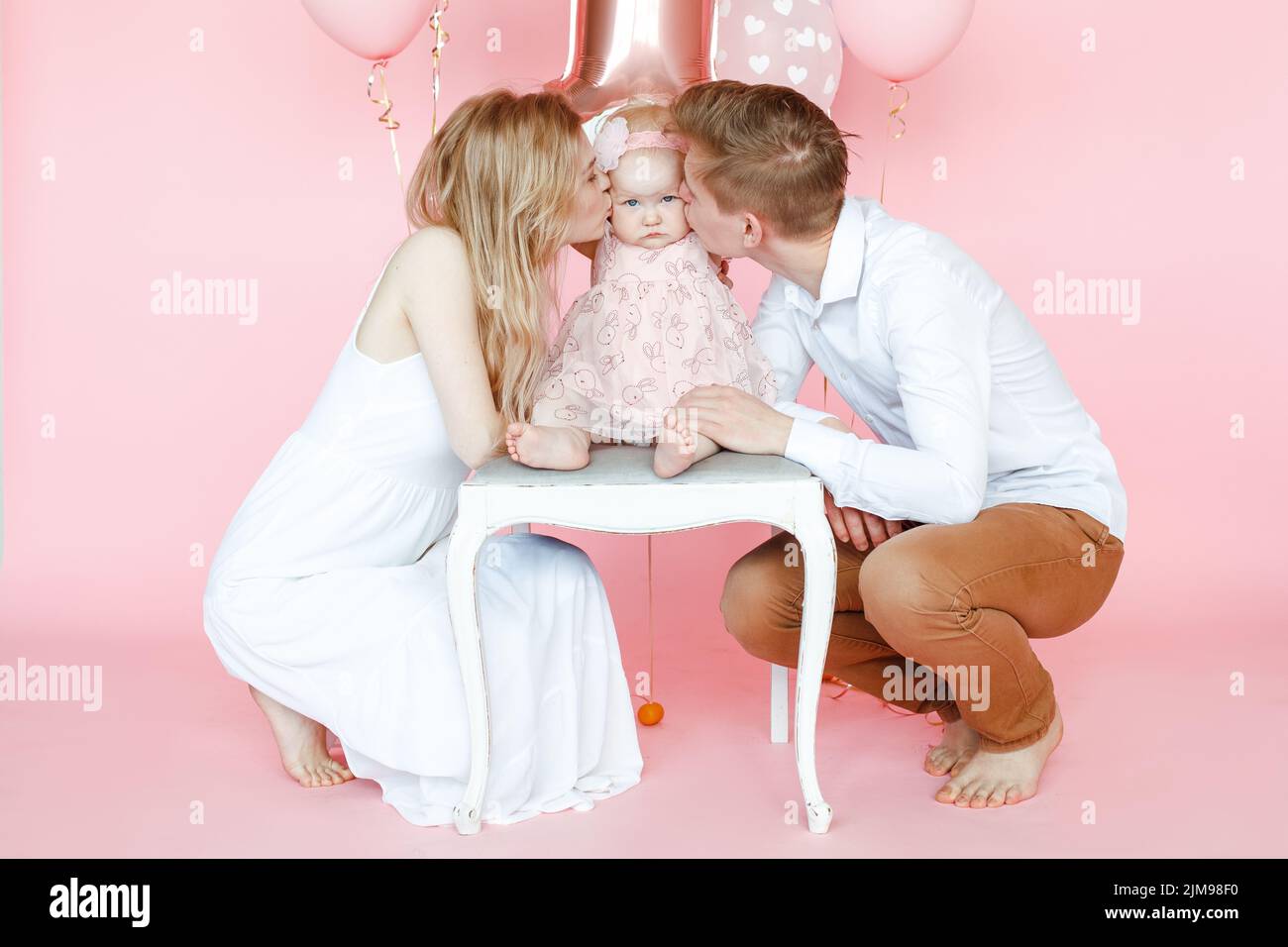 Calm, loving full family of man, woman kissing one year infant in pink dress sit on chair and celebrate birthday party Stock Photo