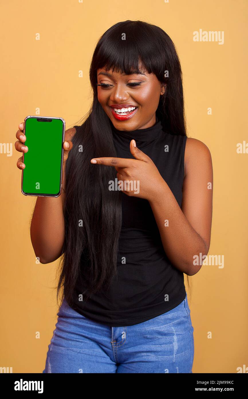 Head shot portrait close up smiling beautiful African American girl showing, pointing finger to green phone screen mock up, demonstrating shopping off Stock Photo