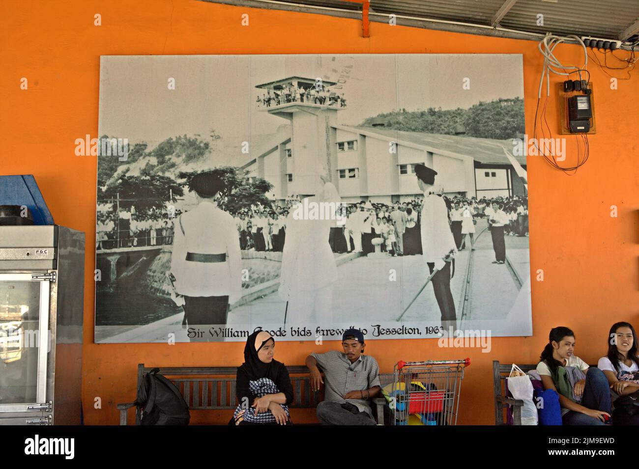 Passengers waiting for transportation as they are sitting in front of a wall where a large photo print of a historical moment is hung at Jesselton Point, a harbour for boats connecting adjacent islands with Kota Kinabalu, which is located in the capital city of Sabah state in Sabah, Malaysia. Stock Photo