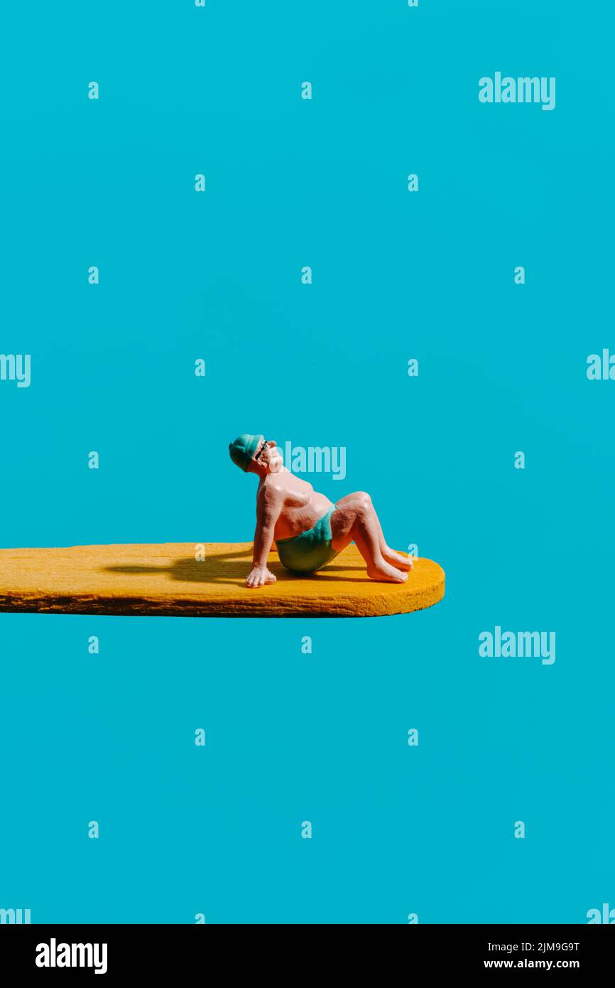 a miniature man, wearing a blue swimsuit, a blue swimming cap and googles, sits on a yellow diving board, on a blue background with some blank space o Stock Photo