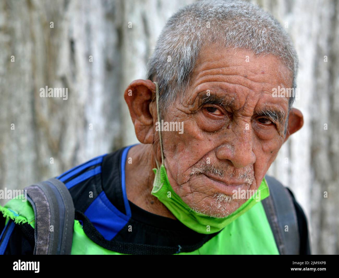 Streetwise poor old Mexican male senior citizen with homemade green fabric face mask under his chin looks silently at the viewer. Stock Photo