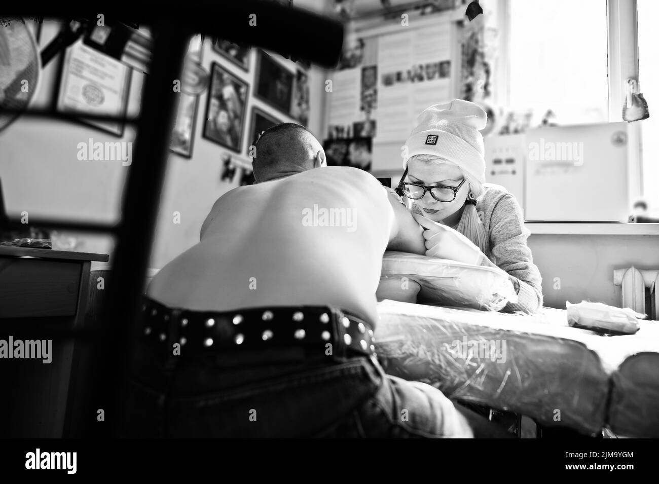 Young blonde woman master tattooist in glasses and hat with tattoo machine at hands make tattoo Dreamcatcher at hand of man. Bla Stock Photo