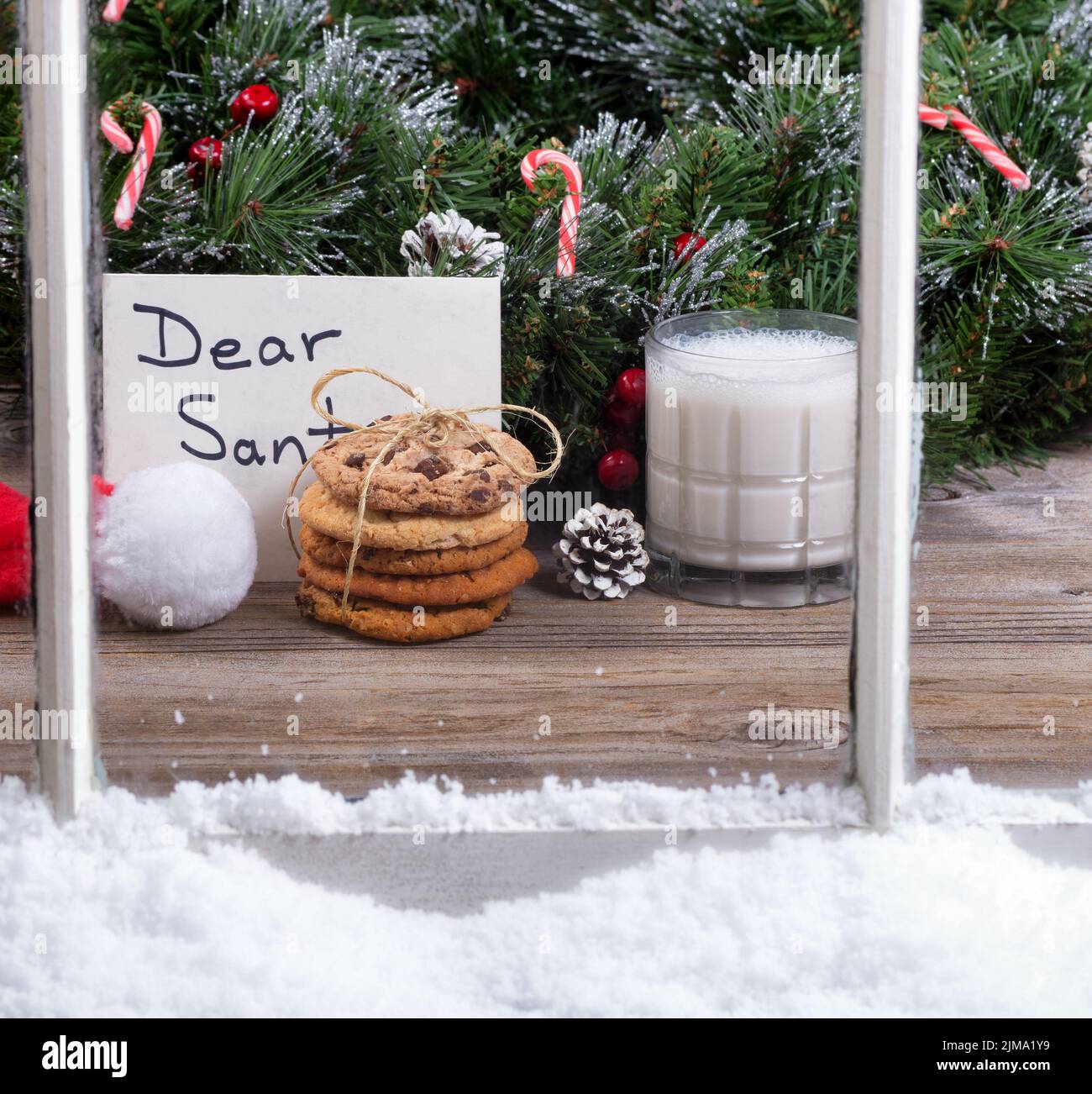 Snowy window view of cookies and milk plus card for Santa Claus with holiday decorations in background Stock Photo