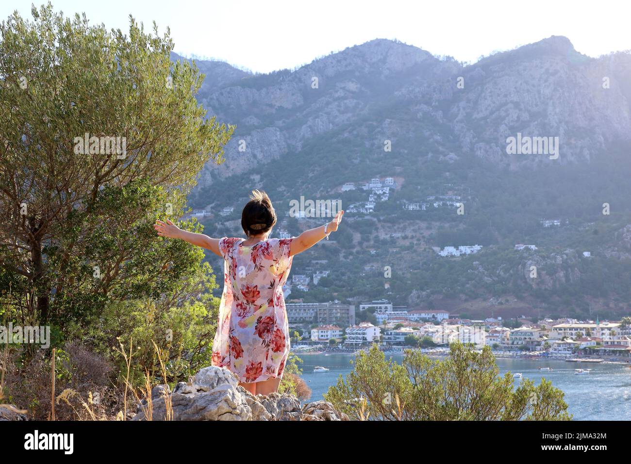Woman in summer dress standing with her arms outstretched on top of the rock in sunbeams and looking to resort town on a beach. View to green mountain Stock Photo