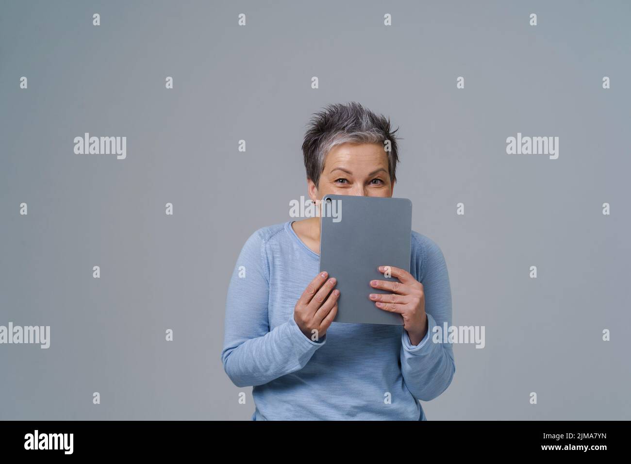 Shy mature woman hide holding digital tablet working or shopping online, checking on social media. Pretty grey haired woman in blue blouse isolated on white background. Stock Photo