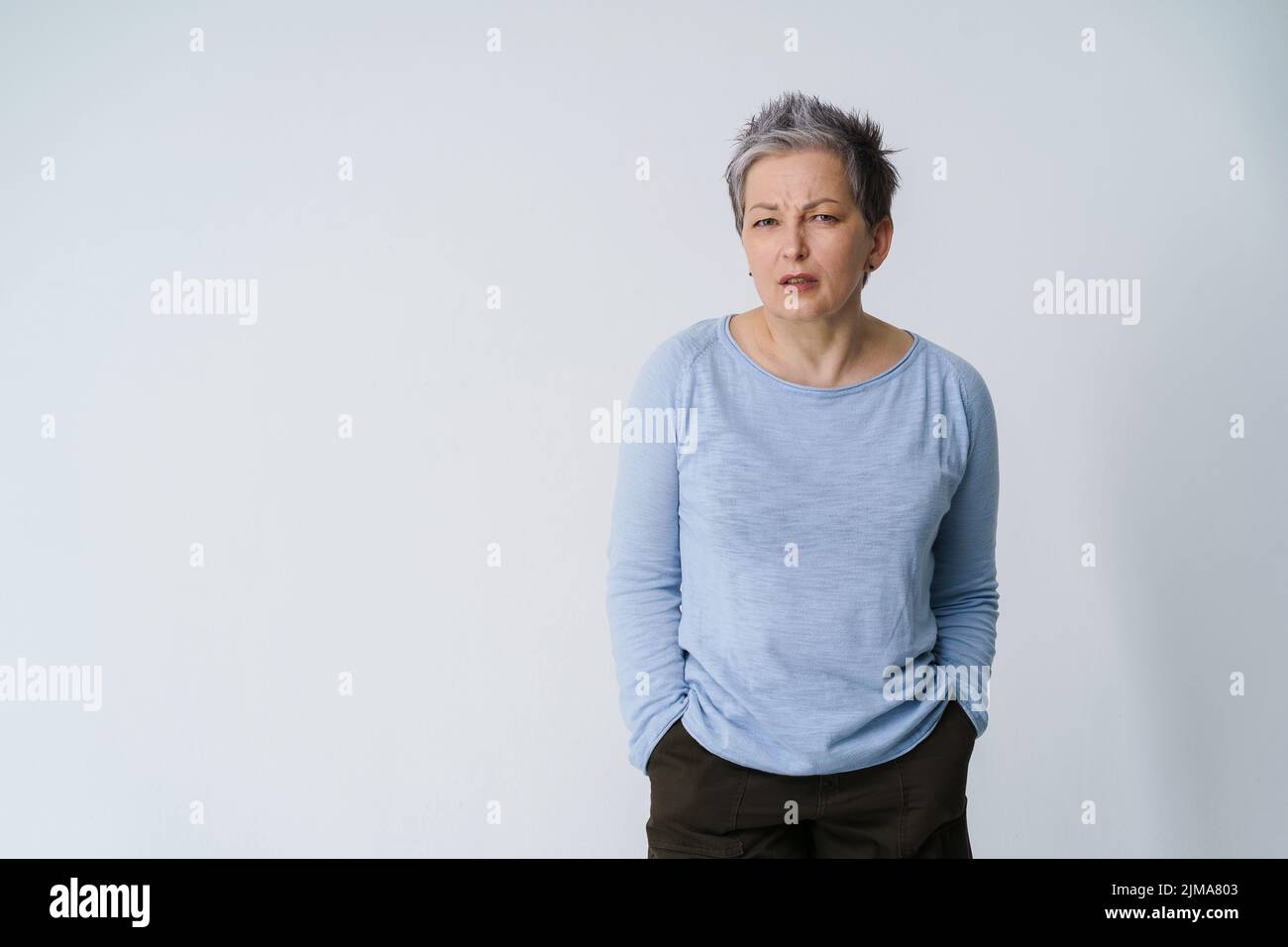 Stressed, angry mature grey haired woman 50s posing frustrated looking at camera with hands in pockets, copy space on left isolated on white background. Mature people healthcare.  Stock Photo