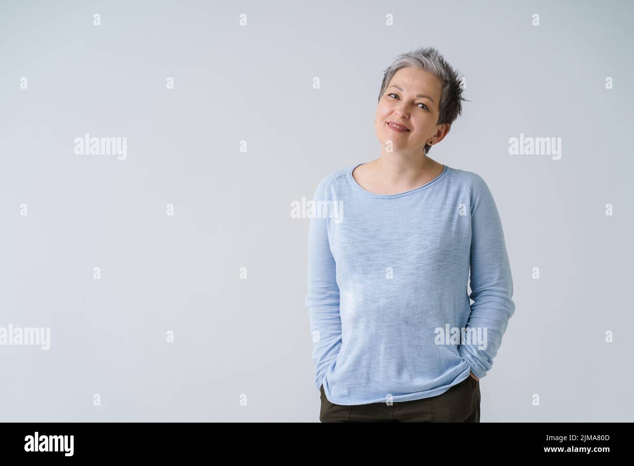 Happy dreamy mature grey haired woman 50s posing tenderly looking at camera with hands in pockets, copy space on left isolated on white background. Mature people healthcare.  Stock Photo