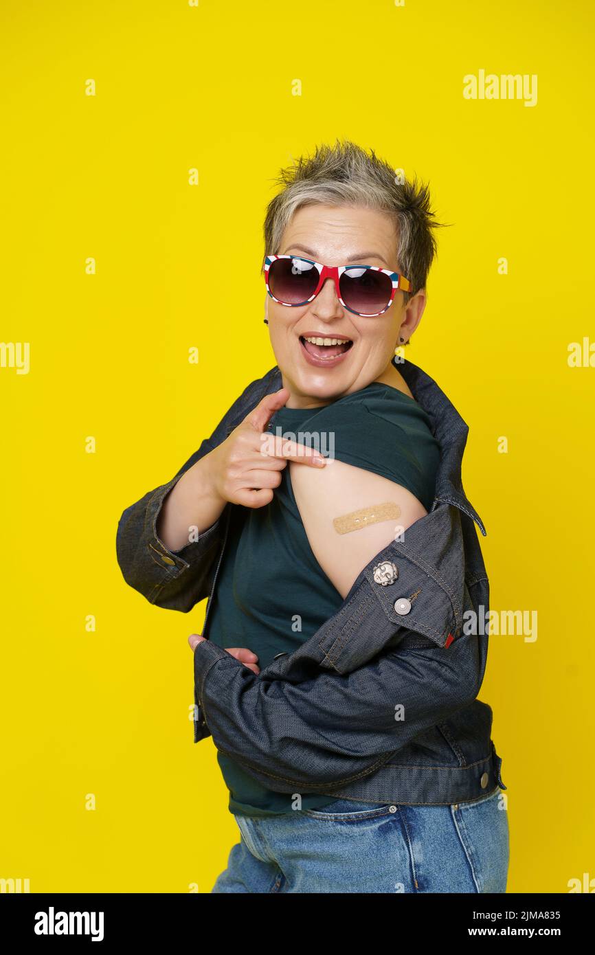 Happy mature grey hair woman showing shoulder with band aid after having shot of vaccine wearing UK flag sunglasses isolated on yellow background. Mature woman 50s vaccination and healthcare concept. Stock Photo