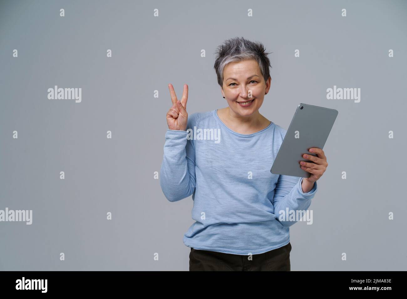 Gesturing V or victory mature grey haired woman in 50s holding digital tablet working or shopping online, checking on social media. Pretty woman in blue blouse isolated on white background. Stock Photo