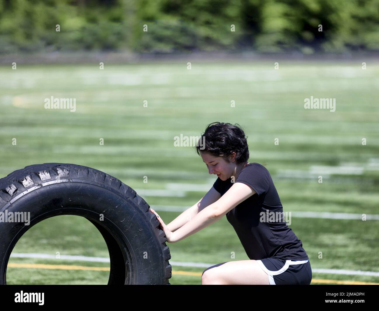 Teen girl pushing heavy old tire on sports field during hot day Stock Photo