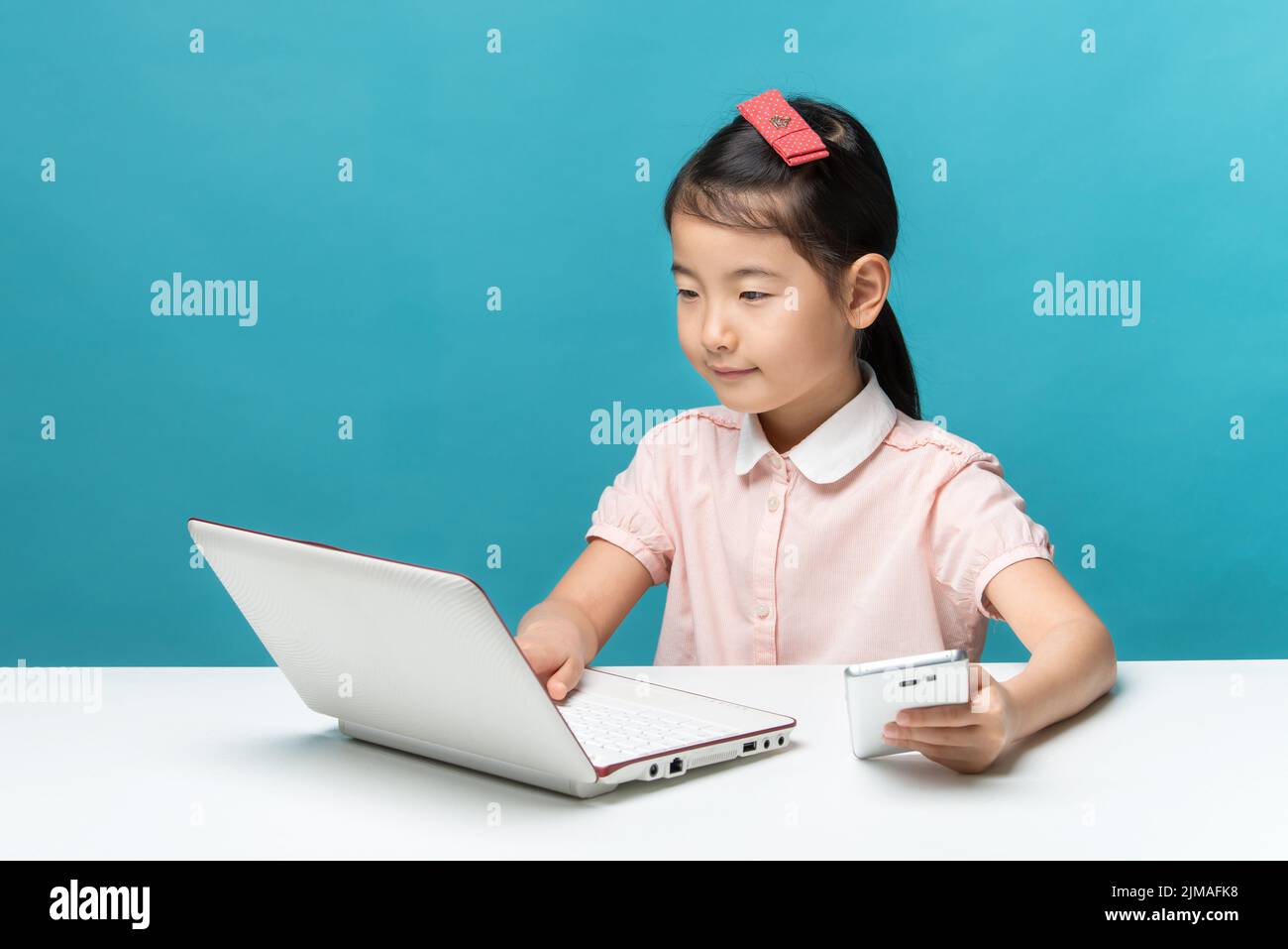 Cute asia little girl is sitting at table with her white laptop and a smartphones, isolated over blu Stock Photo
