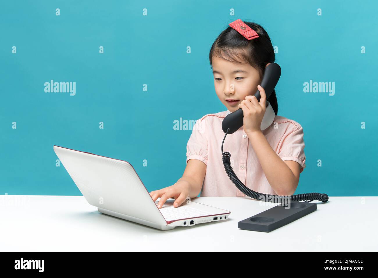 Cute asia little girl is sitting at table with her white laptop and a telephone, isolated over blue Stock Photo