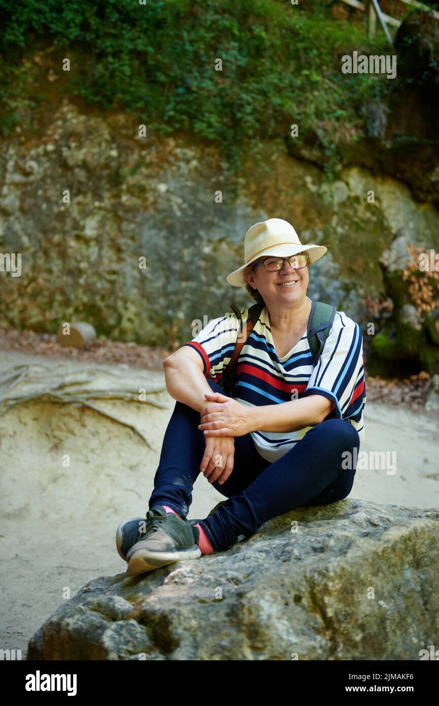 Middle-aged woman with hat smiling sitting and resting on a rock from the path in a natural environment, Urederra, Baquedano, Navarra, Spain, Europe Stock Photo