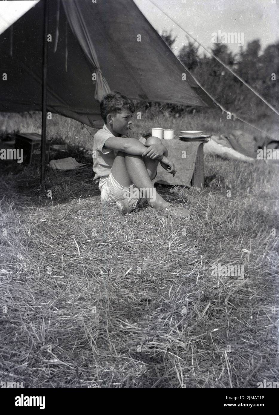 1930s, historical, summertime and a young boy sitting on the grass in a field underneath a canvas tent canopy, England, UK. Stock Photo