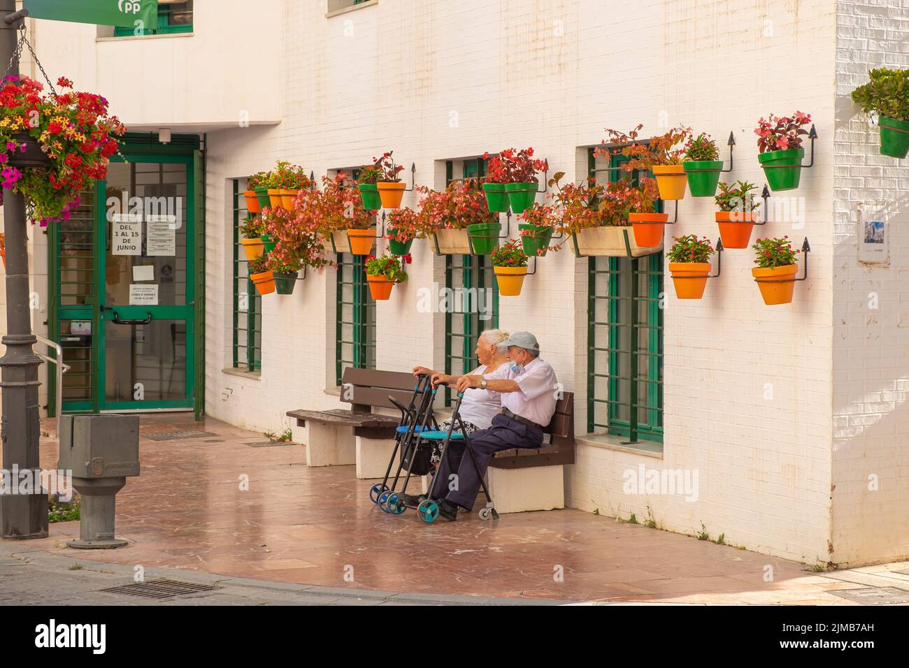Elderly couple sitting on a bench in in a pedestrian street full of colorful flowers in pots, typical of Estepona. Old couple, life in retirement Stock Photo