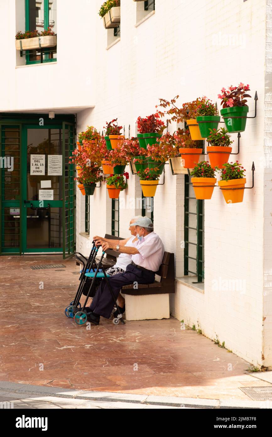 Elderly couple sitting on a bench in in a pedestrian street full of colorful flowers in pots, typical of Estepona. Old couple, life in retiremen Stock Photo