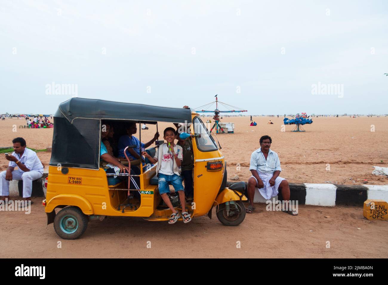 A boy sitting in a touk touk smiles to the camera as other people are enjoying themselves on Marina Beach in Chennai. Stock Photo