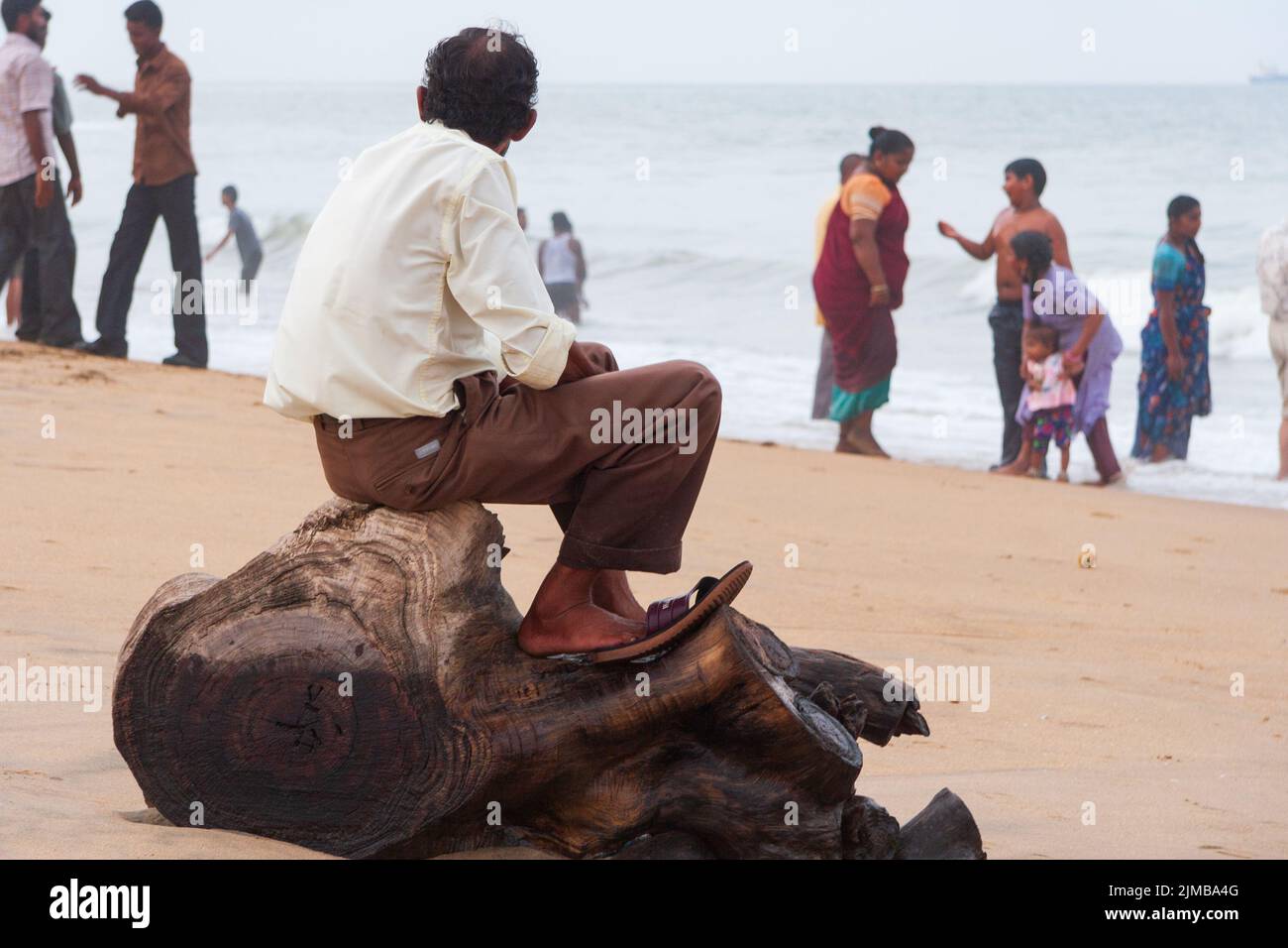 A man sitting on a tree stump gazes towards the rough sea as other people enjoy themselves at Marina Beach in Chennai. Stock Photo