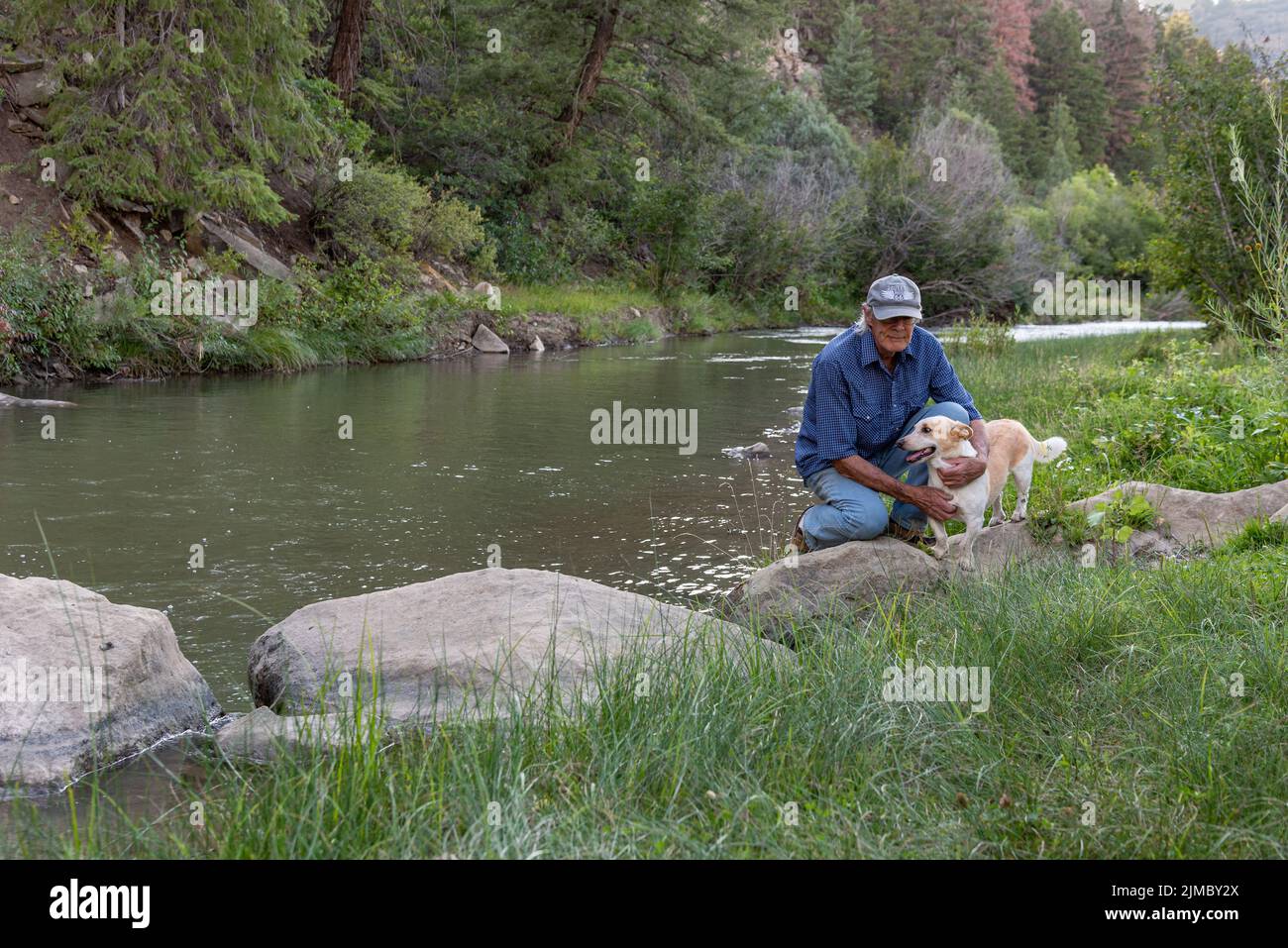 A senior male crouches on a boulder next to his dog, a corgi mix, by the Navajo River in Dulce, New Mexico. Stock Photo
